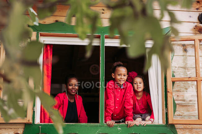 Positive young black woman with kids dressed in red clothes looking at camera while standing near open window of wooden rural house — Stock Photo
