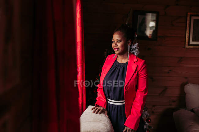 Vista laterale di positiva femmina nera in piedi vicino alla finestra in accogliente casa di legno con albero di Natale — Foto stock