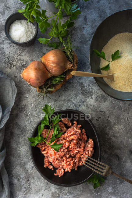 D'en haut vue oignons crus et herbes placées sur la table grise près du bol avec du sel de viande hachée dans la cuisine — Photo de stock