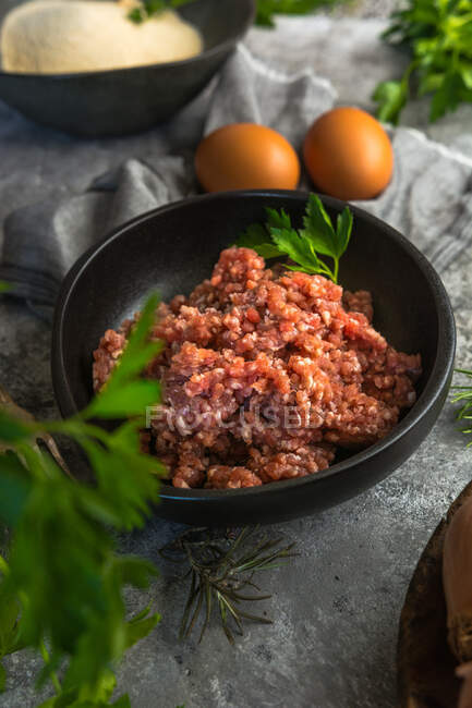 From above bowl of fresh minced meat placed on gray surface near green herbs and chicken eggs in kitchen — Stock Photo