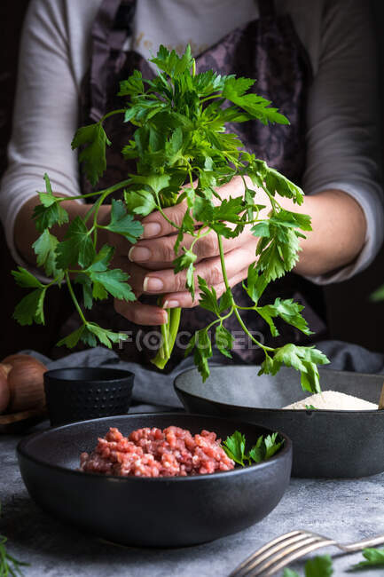 Unrecognizable female wearing apron keeping bunch of fresh parsley over bowl with raw minced meat while cooking lunch in kitchen — Stock Photo