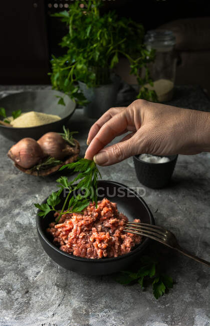 Unrecognizable person putting fresh parsley into bowl with raw minced meat while preparing lunch — Stock Photo