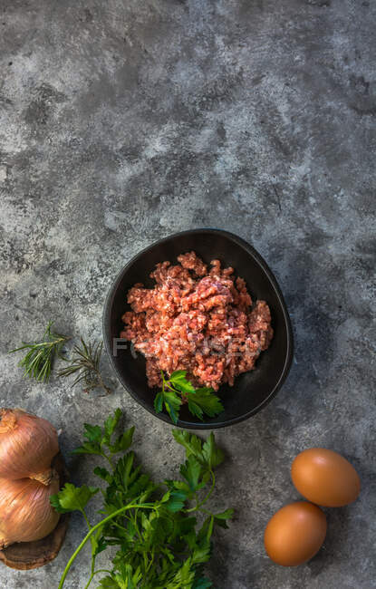 De cima vista superior cebolas cruas, ovos e ervas colocadas em mesa cinza perto tigela com sal de carne picada na cozinha — Fotografia de Stock