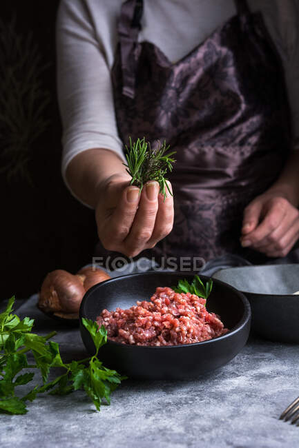 Cropped unrecognizable female wearing apron keeping bunch of fresh leaves over bowl with raw minced meat while cooking lunch in kitchen — Stock Photo
