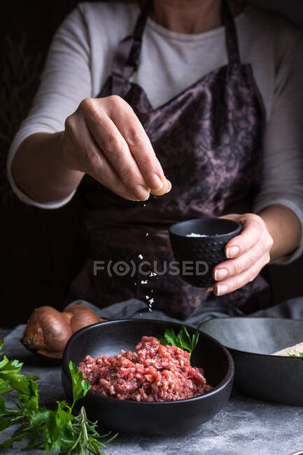 Anonymous female in apron spilling salt on fresh minced meat while preparing lunch in kitchen — Stock Photo