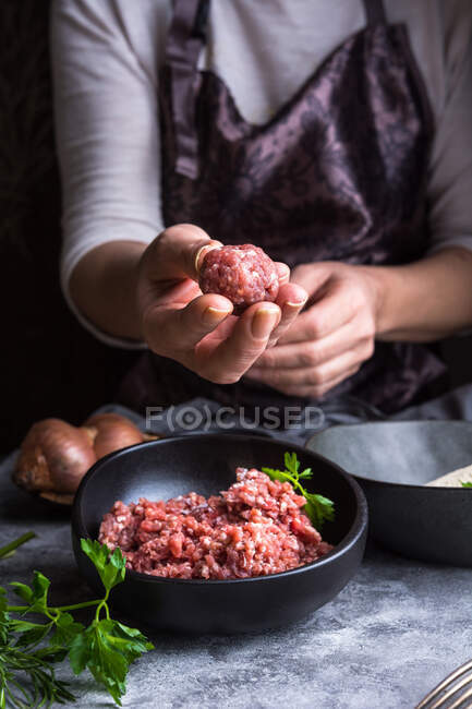 From above unrecognizable person wearing apron showing meatball to camera over bowls with minced meat during lunch preparation — Stock Photo