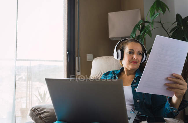Mulher madura ouvindo música em fones de ouvido e lendo papel enquanto sentada em poltrona e fazendo trabalho remoto no laptop perto da janela em casa — Fotografia de Stock