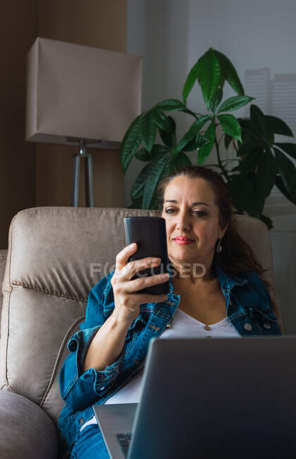 Зрелая женщина с ноутбуком просматривает смартфон и смотрит в сторону, сидя в кресле и работая над удаленным проектом дома — стоковое фото