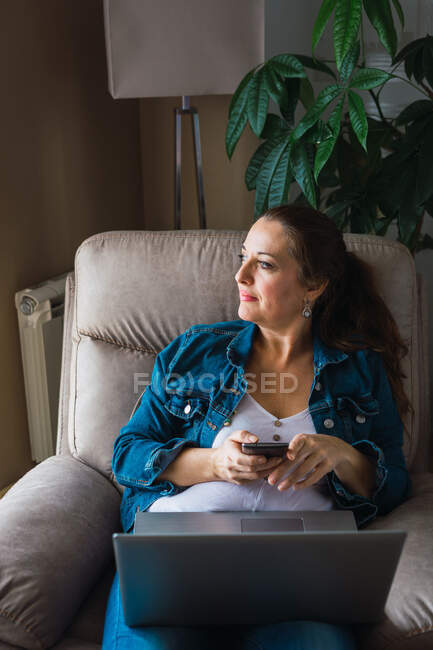Зріла жінка з ноутбуком переглядає смартфон і дивиться вбік, сидячи в кріслі і працюючи на віддаленому проекті вдома — стокове фото