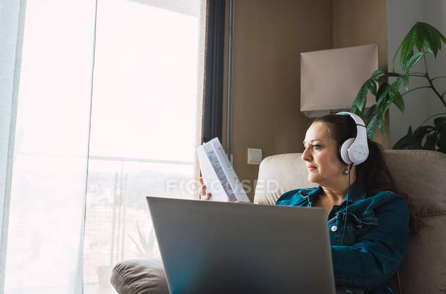 Зріла жінка, яка слухає музику в навушниках і читає газету, сидячи в кріслі і роблячи дистанційну роботу на ноутбуці біля вікна вдома. — стокове фото