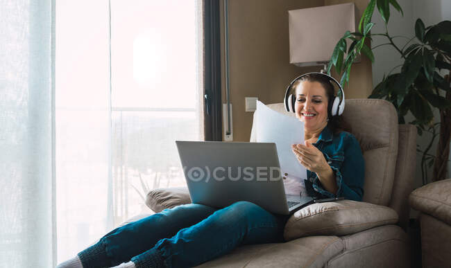Donna matura che ascolta musica in cuffia e legge carta mentre è seduta in poltrona e fa un lavoro remoto sul computer portatile vicino alla finestra di casa — Foto stock