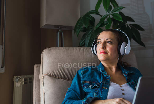Зріла жінка слухає музику в навушниках, сидячи в кріслі і роблячи віддалену роботу на ноутбуці біля вікна вдома, дивлячись далеко — стокове фото