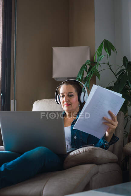 Зріла жінка слухає музику в навушниках і читає папір, сидячи в кріслі і роблячи віддалену роботу на ноутбуці біля вікна вдома — стокове фото