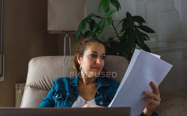 Зрелая женщина читает бумагу, сидя в кресле и выполняя удаленную работу на ноутбуке у окна дома — стоковое фото