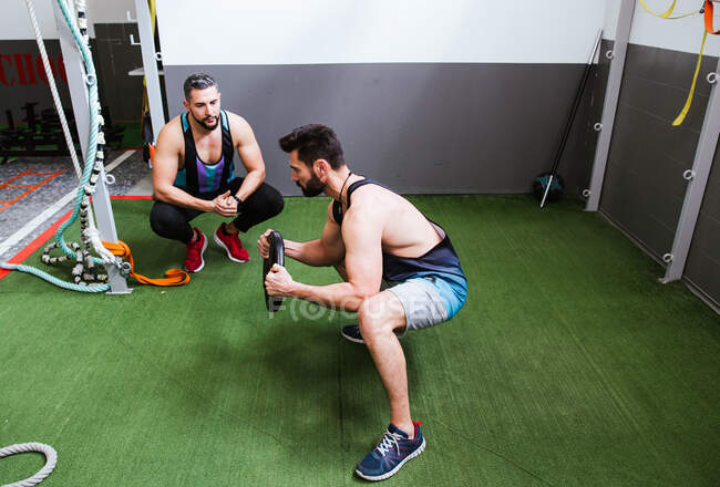 Hochwinkelseitenansicht des fokussierten starken Sportlers beim Kniebeugen mit der Gewichtsscheibe während des Trainings im Fitnessstudio mit dem Trainer — Stockfoto