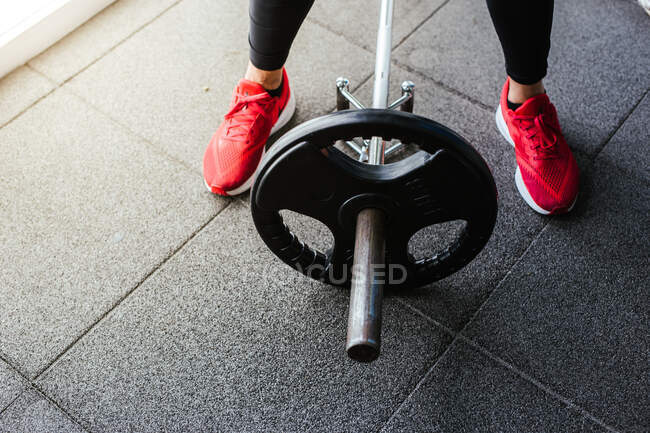 From above of crop anonymous athlete in black leggings and red sneakers standing near barbell with weight in gym — Stock Photo