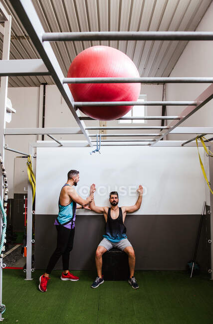 Jeune homme athlétique en vêtements de sport effectuant des squats tout en s'entraînant dans une salle de gym moderne avec instructeur — Photo de stock