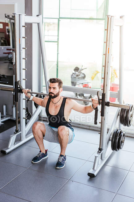Мощный целеустремленный спортсмен делает упражнения с штангой на корточках во время тренировки тяжелой атлетики с личным тренером в современном тренажерном зале — стоковое фото