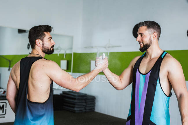 Vista lateral de musculosos culturistas masculinos competitivos con palmas de talco en polvo saludándose entre sí mientras están de pie en el gimnasio moderno - foto de stock