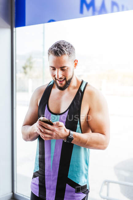 Возбужденный мускулистый мужчина в спортивной одежде, сосредоточенный на экране и с удовольствием взаимодействующий со смартфоном, стоя в коридоре современного спортзала напротив стеклянной стены — стоковое фото