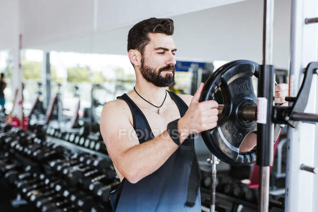 Muskulöser bärtiger Mann in Sportbekleidung, der in der Nähe eines Kniegelenks steht und die Hantelscheibe auf die Langhantel legt, während er sich auf das Gewichtheben in einem modernen Fitnessstudio vorbereitet — Stockfoto
