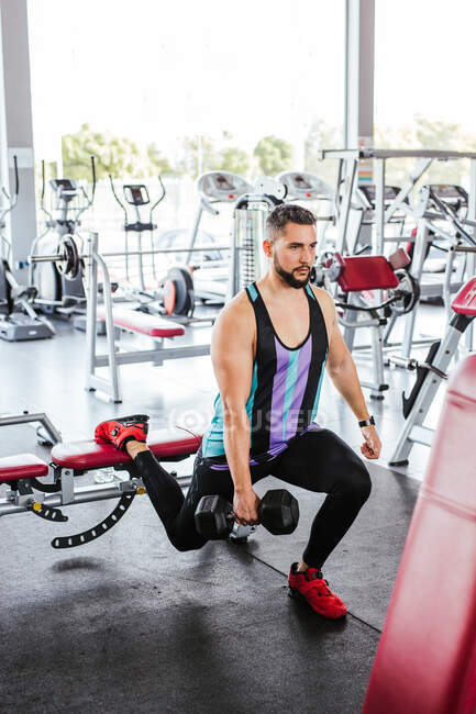 High angle of calm confident adult man in activewear performing single leg squat exercise with dumbbell while leaning on bench against blurred interior and sports equipment in modern spacious fitness club — Stock Photo