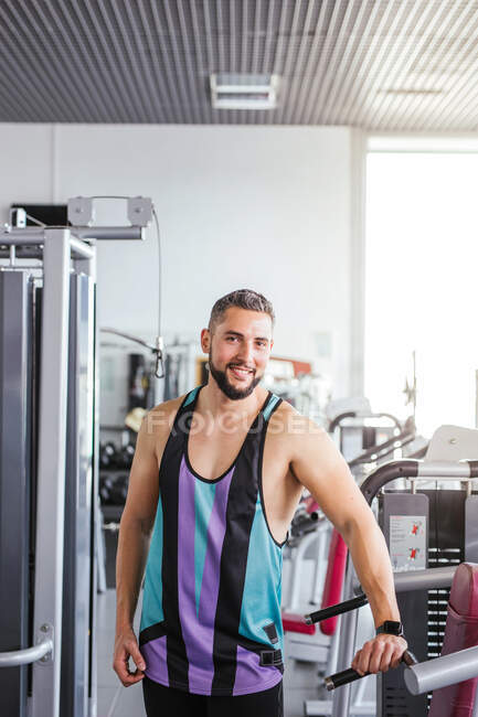 Muscular bearded man in sportswear looking at camera while standing with arms crossed and leaning on sports equipment in light contemporary gym — Stock Photo