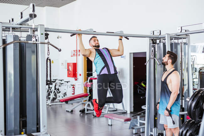 Low angle of pensive instructor watching determined athlete in sportswear performing pull ups exercise on bar in contemporary sport club — Stock Photo