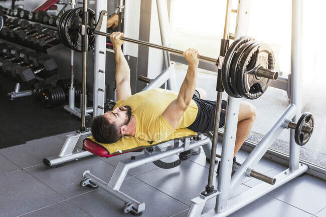 High angle of calm sportsman in activewear performing exercise with barbell while laying on bench near window wall in modern sports center — Stock Photo