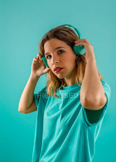 Side view of thoughtful young woman in bright t shirt looking at camera while listening to music in headphones against turquoise background — Stock Photo