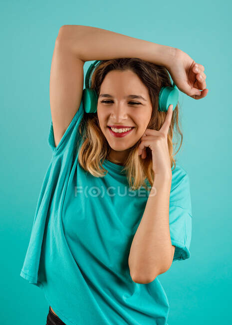 Mujer joven feliz en camiseta brillante sonriendo y escuchando música en auriculares mirando hacia otro lado sobre fondo turquesa - foto de stock