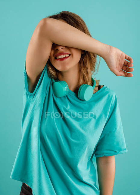 Felice giovane donna in t shirt luminosa sorridente con il braccio sul viso che copre gli occhi con le cuffie poggiate nel collo contro lo sfondo turchese — Foto stock