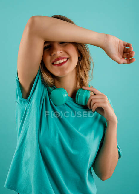 Mujer joven feliz en camiseta brillante sonriendo mirando a la cámara con el brazo sobre la frente con auriculares descansando en su cuello sobre fondo turquesa - foto de stock