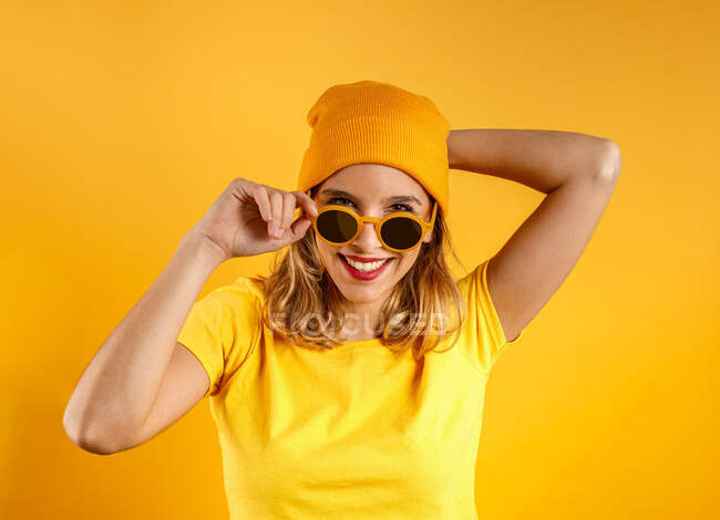 Felice giovane donna in abiti luminosi e occhiali da sole alla moda sorridente e guardando la fotocamera sullo sfondo arancione — Foto stock