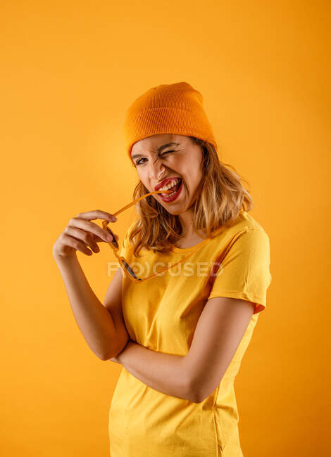 Вид сбоку счастливой молодой женщины в яркой одежде и солнцезащитных очках, улыбающейся и смотрящей на камеру, моргая глазами на оранжевом фоне — стоковое фото