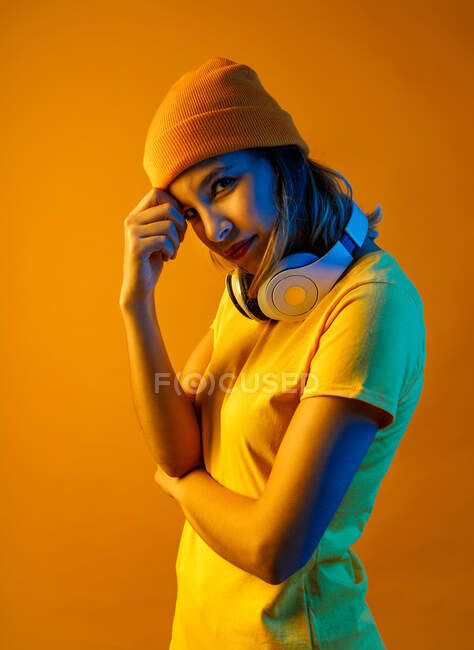 Mujer joven con estilo reflexivo en gorro naranja con auriculares alrededor del cuello descansando cabeza sobre brazo y mirando a la cámara contra fondo naranja - foto de stock