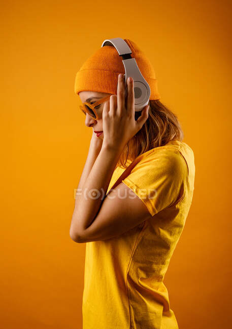 Щаслива молода жінка в яскравому одязі, що регулює стильні сонцезахисні окуляри і посміхається, дивлячись в сторону, слухаючи музику на помаранчевому фоні — стокове фото