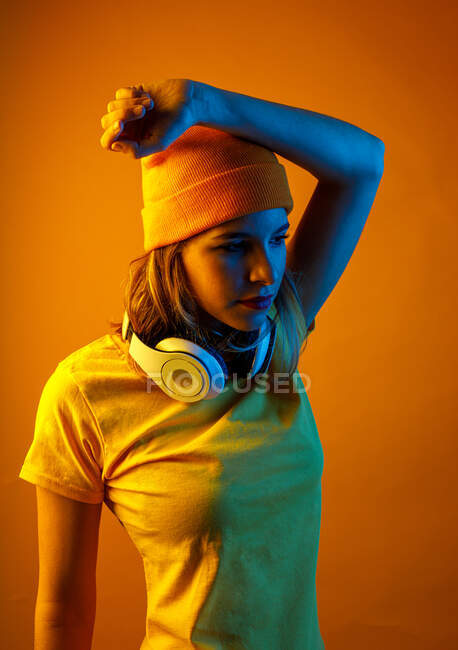 Продумана молода жінка в помаранчевій бляшанці з навушниками навколо шиї тримає руку над головою і дивиться на помаранчевий фон — стокове фото