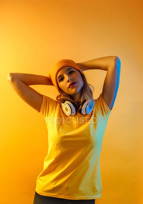 Selbstbewusste stilvolle Frau mit Kopfhörern, die die Hände hinter dem Kopf hält und vor orangefarbenem Hintergrund in die Kamera blickt — Stockfoto