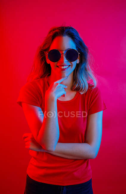 Donna positiva in occhiali da sole rotondi toccare mento e sorridente per la fotocamera mentre in piedi sotto la luce blu sullo sfondo rosso — Foto stock