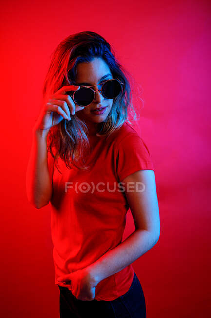 Thoughtful young blonde woman in round sunglasses looking away while standing against red background — Stock Photo