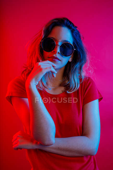Nachdenkliche junge blonde Frau mit runder Sonnenbrille, die das Kinn berührt und vor rotem Hintergrund in die Kamera blickt — Stockfoto
