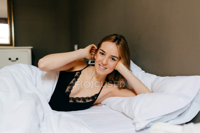 Досить молода жінка в нижній білизні лежить в ліжку посміхаючись і дивлячись на камеру вранці в затишній спальні вдома — стокове фото