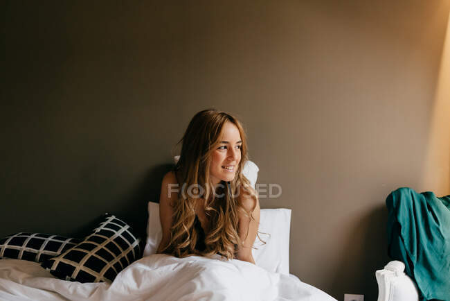 Jolie jeune femme en sous-vêtements assis dans le lit souriant et regardant loin le matin dans une chambre confortable à la maison — Photo de stock