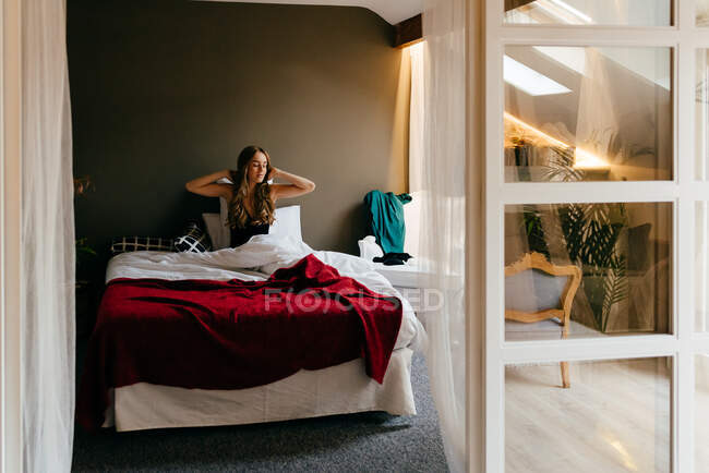 Feliz serena jovem fêmea em sleepwear levantando braços com os olhos fechados esticando o corpo enquanto sentado em cama confortável depois de acordar de manhã em casa — Fotografia de Stock