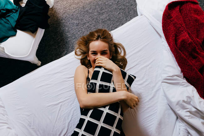 Vista dall'alto di eccitata bella giovane femmina in biancheria intima ridendo guardando la fotocamera e gettando cuscino mentre sdraiato su un comodo letto al mattino a casa — Foto stock
