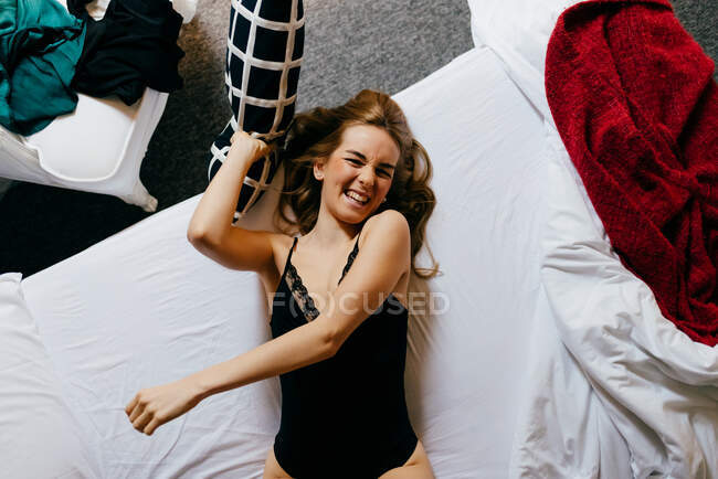 Вид зверху збуджена красива молода жінка в нижній білизні сміється, дивлячись на камеру і кидаючи подушку, лежачи на зручному ліжку вранці вдома — стокове фото