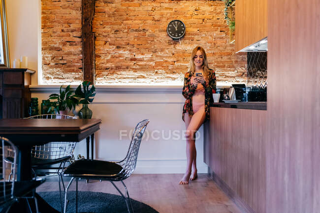 Чувственная молодая блондинка в нижнем белье и цветочная блузка, стоящая утром на кухне и просматривающая смартфон — стоковое фото