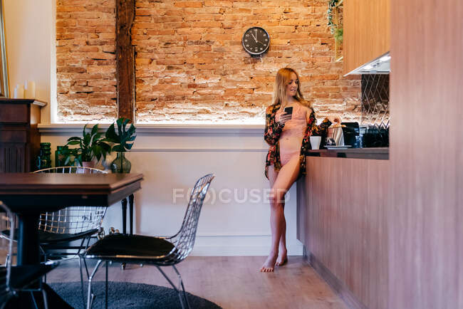 Чуттєва блондинка в нижній білизні і квіткова блузка, що стоїть на кухні, готує тости на сніданок під час перегляду на смартфоні — стокове фото