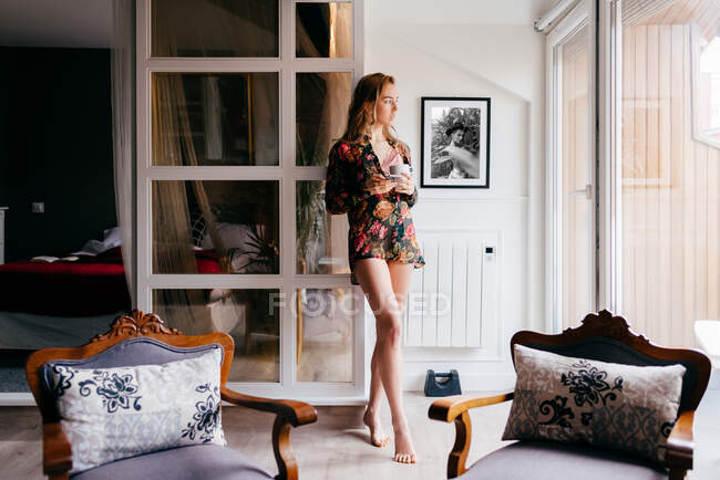 Jolie mince réfléchie jeune femme en robe de soie colorée boire du café en regardant loin tout en se tenant devant la fenêtre dans le salon confortable le matin — Photo de stock